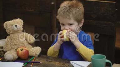 可爱的金发男孩坐在木椅上，在木制背景上吃大红苹果。 学校休息。 饥饿的孩子